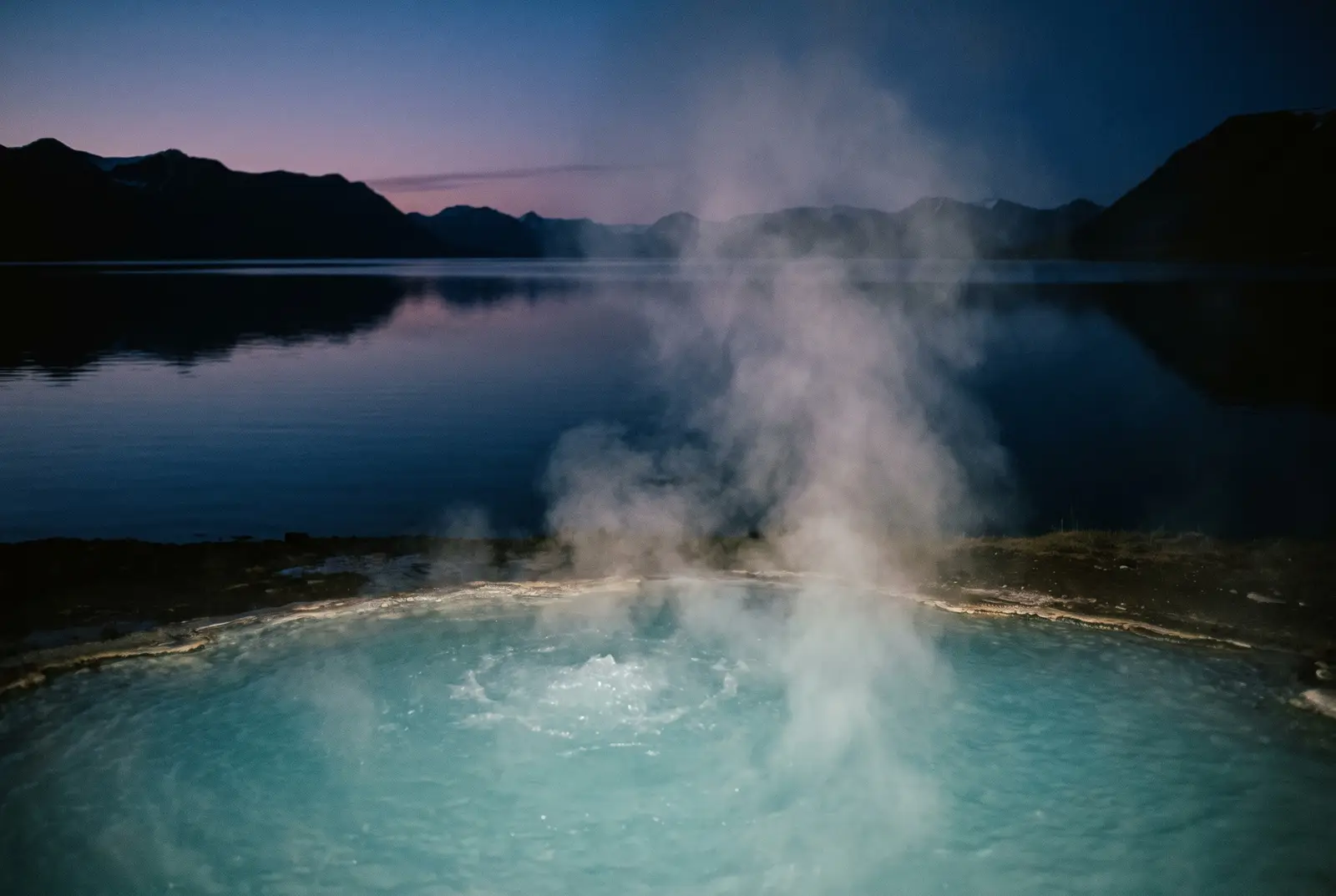Vapor elevándose de una poza geotermal turquesa junto a un lago espejo en los fiordos del este